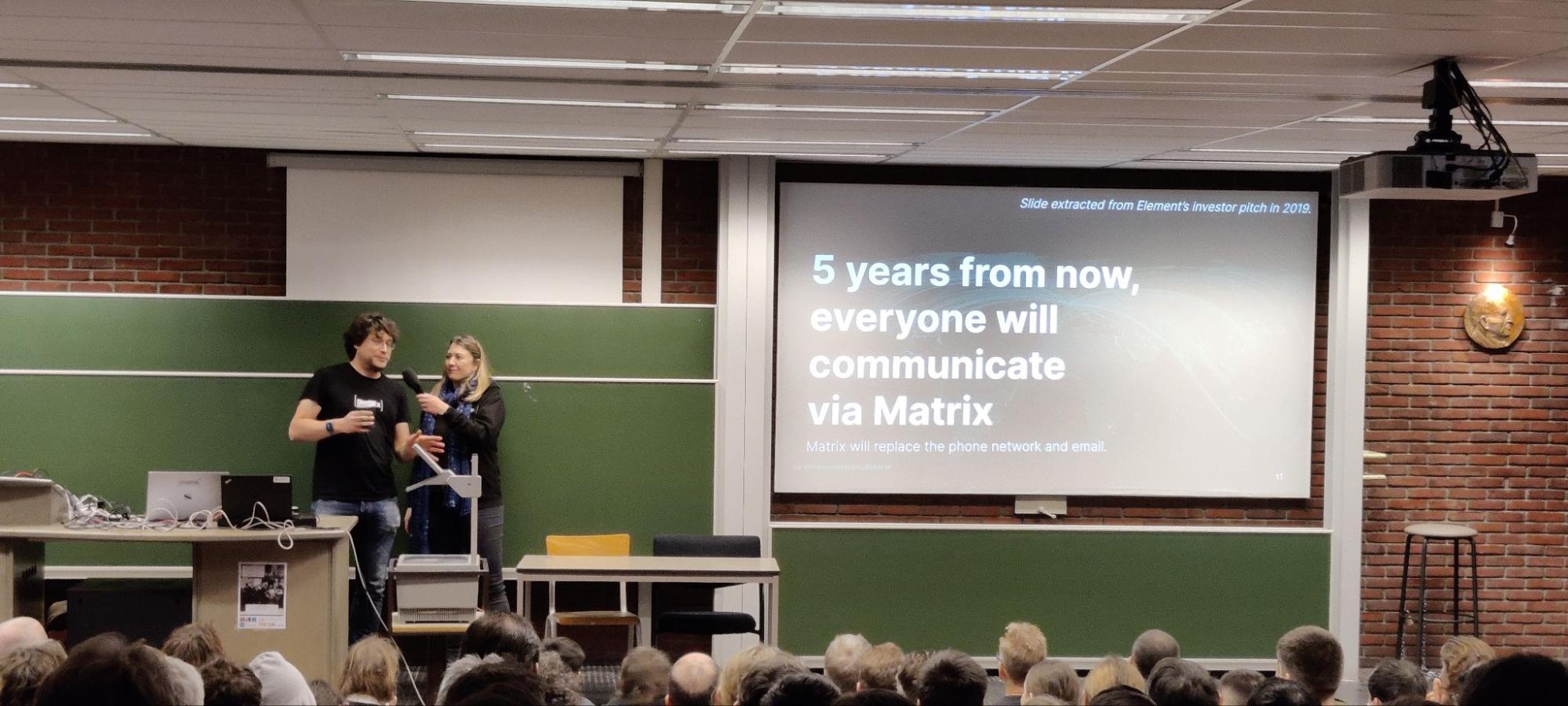 A picture of Matthew and Amandine in front of a classroom with a wide audience. Amandine is holding the mic for Matthew. Their slides are projected, and we can ready "5 years from now, everyone will communicate via Matrix"