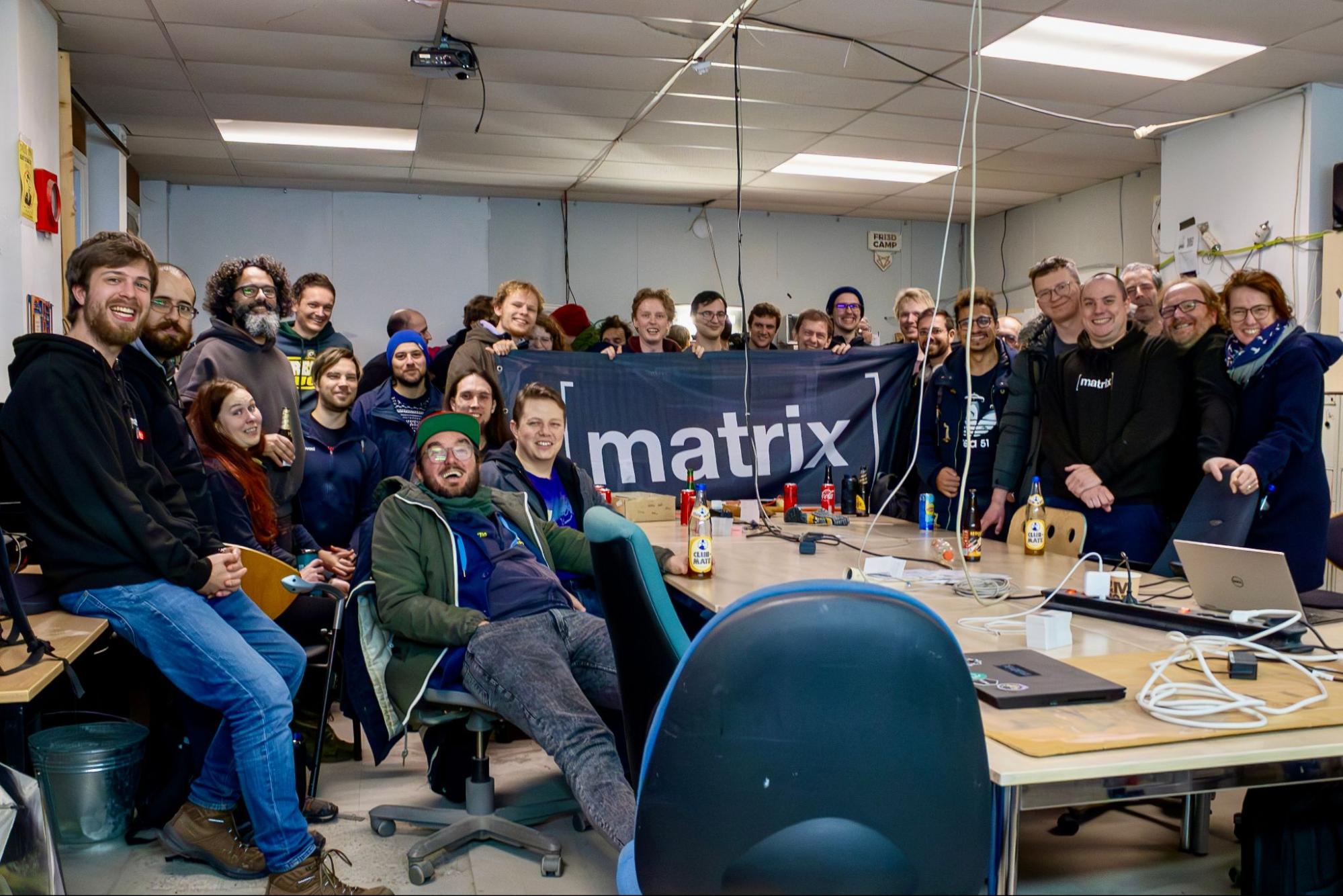 A picture of several dozens of people smiling in a room. Some of them are holding a Matrix flag together.